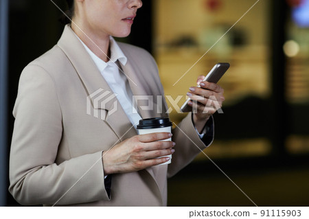 Close-up of pensive woman in jacket drinking coffee from takeout cup and reading sms on phone Close-up of pensive woman in jacket drinking coffee from takeout cup and reading sms on phone 91115903
