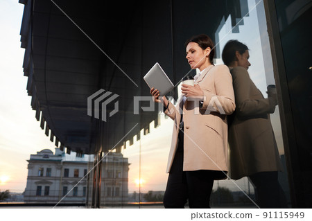 Below view of pensive attractive middle-aged lady in elegant jacket leaning on building wall and watching video presentation on tablet Below view of pensive attractive middle-aged lady in elegant jacket leaning on building wall and watching video presentation on tablet 91115949
