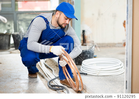 Qualified builder in blue workwear plans to make warm floor, prepares plastic pipes Qualified builder in blue workwear plans to make warm floor, prepares plastic pipes 91120295