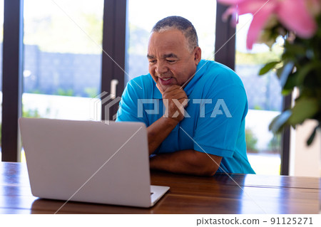 Smiling biracial senior man with hand on chin watching video over laptop while sitting at table 91125271