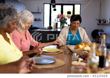 Multiracial senior female friends holding hands and saying grace at dining table in nursing home Multiracial senior female friends holding hands and saying grace at dining table in nursing home 91125429