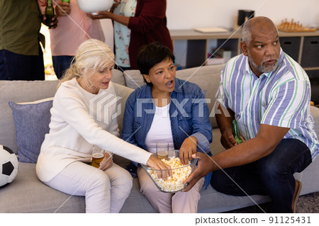Multiracial senior friends with beer having popcorn while watching soccer match in nursing home 91125431