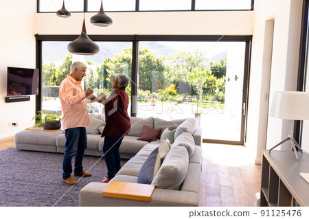 Happy multiracial senior friends holding hands and dancing by sofa in nursing home 91125476