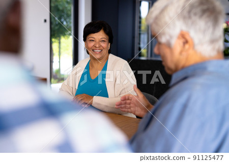 Smiling biracial senior woman talking with male friend while sitting at dining table in nursing home 91125477