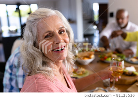 Portrait of senior multiracial woman enjoying wine while having lunch with friends at dining table 91125536