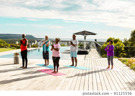 Multiracial seniors meditating while standing on hardwood floor at poolside against sky in summer Multiracial seniors meditating while standing on hardwood floor at poolside against sky in summer 91125556