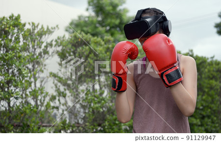 Young female wearing VR headset for working out boxing, aerobic training for boxing punch in virtual reality in simulation world. Young female wearing VR headset for working out boxing, aerobic training for boxing punch in virtual reality in simulation world. 91129947