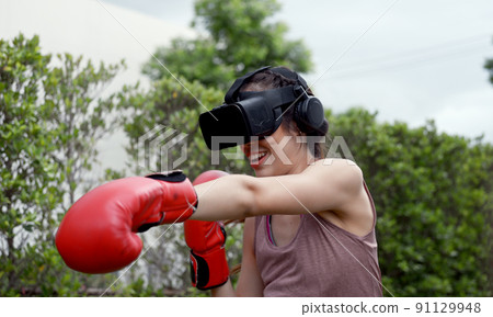 Young female wearing VR headset for working out boxing, aerobic training for boxing punch in virtual reality in simulation world. Young female wearing VR headset for working out boxing, aerobic training for boxing punch in virtual reality in simulation world. 91129948