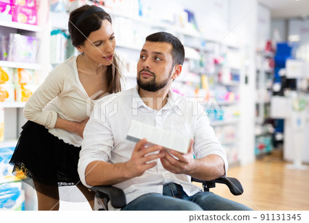 Courteous woman helping man in a wheelchair to choose the right medicine in pharmacy 91131345