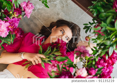 an attractive young woman in a pink blouse with peony flowers on the floor. an attractive young woman in a pink blouse with peony flowers on the floor. 91135219