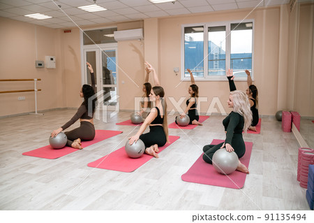 A group of six athletic women doing pilates or yoga on pink mats in front of a window in a beige loft studio interior. Teamwork, good mood and healthy lifestyle concept. A group of six athletic women doing pilates or yoga on pink mats in front of a window in a beige loft studio interior. Teamwork, good mood and healthy lifestyle concept. 91135494