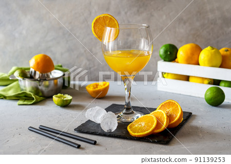 Yellow bird cocktail with rum, orange and lime juice in glass. Fruits in box, juicer, ice cubes, stone board on concrete background 91139253