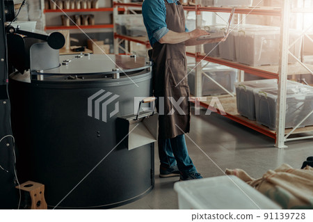 Close up of man on coffee factory working laptop on background of coffee roasting machine 91139728