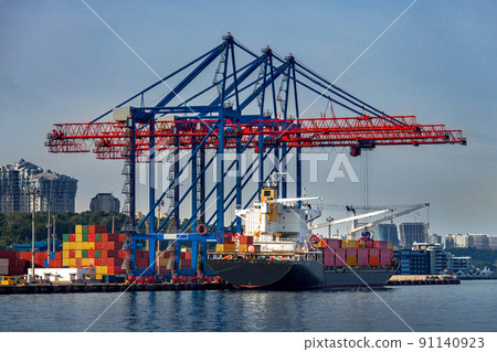 cargo ship moored for loading at container yard with container handling gantry crane or ship-to-shore crane unloading goods from deck to cargo port area, background cityscape with high-rise building. 91140923