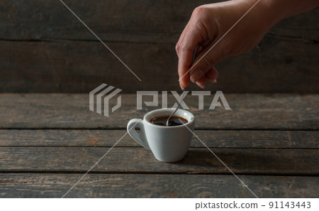 Hot black coffee or espresso in a white ceramic cup on a background of wooden texture stirs a female hand with a spoon Hot black coffee or espresso in a white ceramic cup on a background of wooden texture stirs a female hand with a spoon 91143443
