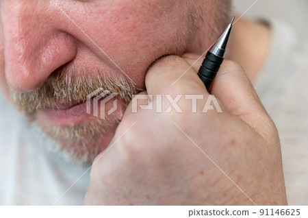 Underside of thoughtful man's face. The man rested his chin on his fist. A black pencil is clenched in his fist. A male with stubble. Gray hair on his beard and mustache. Close-up. Selective focus. 91146625