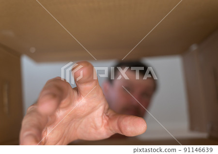 A man's hand reaches into a cardboard box. A middle-aged man is trying to get something out of a large donation box with his hand. Bottom view. Inside view. Close-up. Selective focus. A man's hand reaches into a cardboard box. A middle-aged man is trying to get something out of a large donation box with his hand. Bottom view. Inside view. Close-up. Selective focus. 91146639