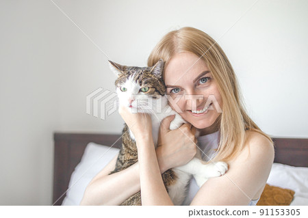 Portrait of young blonde happy 30s woman holding cute tabby cat with green eyes. Female hugging cat in hands 91153305