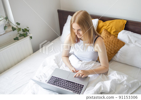 Cropped shot of young woman in pajamas sitting on a bed, using laptop at the morning. Working at home, freelance concept. Cropped shot of young woman in pajamas sitting on a bed, using laptop at the morning. Working at home, freelance concept. 91153306