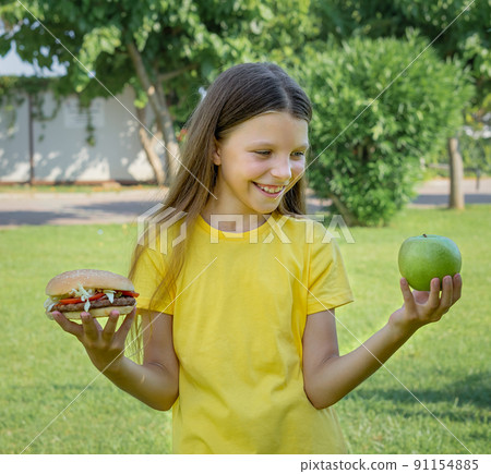 A teenage girl chooses between a hamburger and fruit outdoors. A teenage girl chooses between a hamburger and fruit outdoors. 91154885
