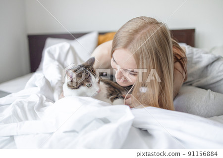 Portrait of young blonde 30s woman holding cute tabby cat with green eyes. Female hugging cat in hands at morning bed Portrait of young blonde 30s woman holding cute tabby cat with green eyes. Female hugging cat in hands at morning bed 91156884