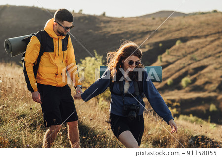 Cheerful Woman with Her Boyfriend Holding by Hands While Hiking on the Hill During Sunset, Young Couple Enjoy Their Trip 91158055
