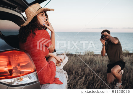 Man Photographing Woman in Red Dress and Hat That Sitting Inside Opened Trunk of the Car on the Background of Sea Man Photographing Woman in Red Dress and Hat That Sitting Inside Opened Trunk of the Car on the Background of Sea 91158056
