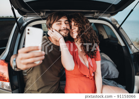 Happy Caucasian Couple Man and Curly Brunette Woman Making Selfie on Smartphone While Sitting in Trunk of the SUV Car Happy Caucasian Couple Man and Curly Brunette Woman Making Selfie on Smartphone While Sitting in Trunk of the SUV Car 91158066