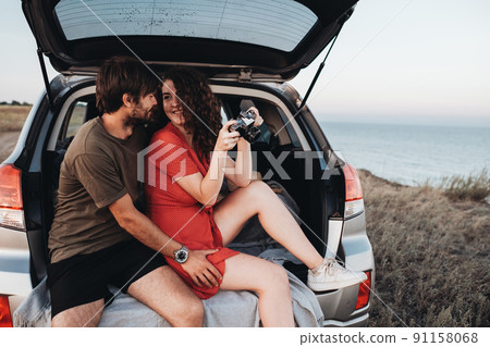 Man and Woman Sitting Inside Opened Trunk of Their SUV Car, Young Couple Enjoying Road Trip Along Sea at Sunset Man and Woman Sitting Inside Opened Trunk of Their SUV Car, Young Couple Enjoying Road Trip Along Sea at Sunset 91158068