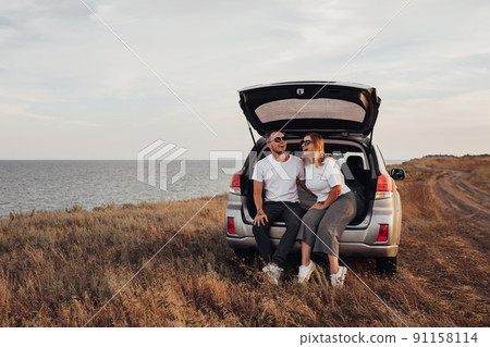 Happy Young Couple Sitting in Open Trunk of the SUV Car, Man and Woman Enjoying Their Road Trip Along Seaside Happy Young Couple Sitting in Open Trunk of the SUV Car, Man and Woman Enjoying Their Road Trip Along Seaside 91158114