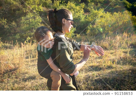A woman looks at a fitness bracelet and uses an ergonomic baby back carrier for a child on a hiking trip A woman looks at a fitness bracelet and uses an ergonomic baby back carrier for a child on a hiking trip 91158653