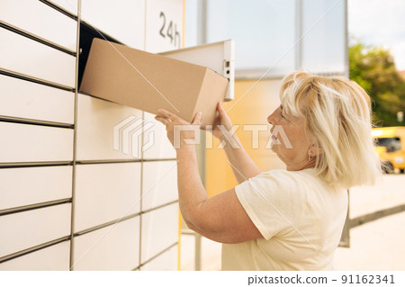 50s caucasian woman getting parcel from cell of automatic post terminal outdoors.  91162341