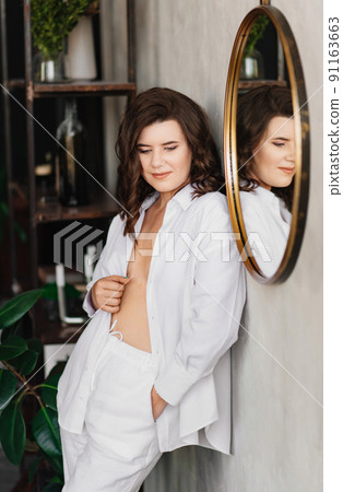 a brunette woman in a white shirt near the round mirror. cozy home clothes. a brunette woman in a white shirt near the round mirror. cozy home clothes. 91163663