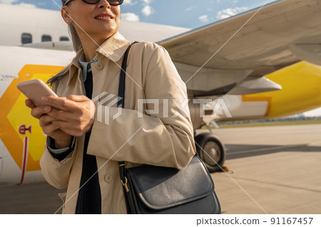 Close up of woman passenger in sunglasses using phone standing outdoors at airport near plane 91167457