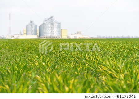 Grain elevator. Metal grain elevator in agricultural zone. Agriculture storage for harvest. Grain elevators on green nature background. Exterior of agricultural factory. Grain elevator. Metal grain elevator in agricultural zone. Agriculture storage for harvest. Grain elevators on green nature background. Exterior of agricultural factory. 91178041