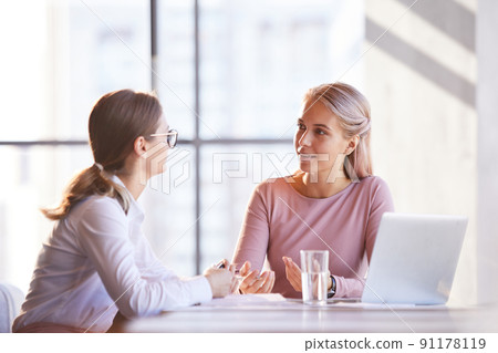 Positive young creative ladies sitting at table with laptop in modern office and sharing ideas for business project 91178119