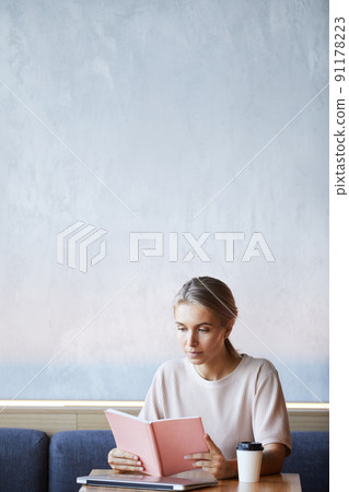 Serious attractive blond woman in sweater sitting at table with laptop and coffee cup and reading book in cafe 91178223
