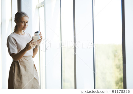 Content attractive blond woman in skirt standing by window and reading message on phone while drinking coffee Content attractive blond woman in skirt standing by window and reading message on phone while drinking coffee 91178236