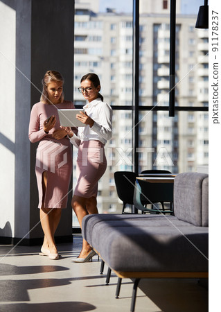 Serious thoughtful young businesswomen in pink clothes standing in modern hall and using tablet while discussing business Serious thoughtful young businesswomen in pink clothes standing in modern hall and using tablet while discussing business 91178237