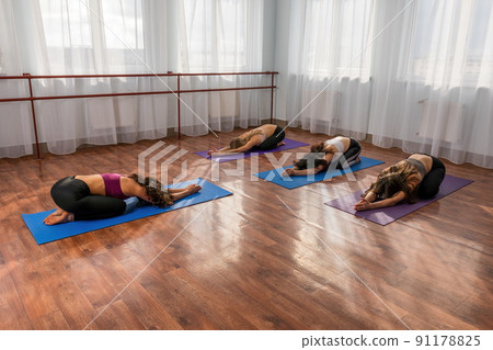 Group of young womans fitness instructor in Sportswear Leggings and Tops, stretching in the gym before pilates, on a yoga mat near the large window on a sunny day, female fitness yoga routine concept 91178825