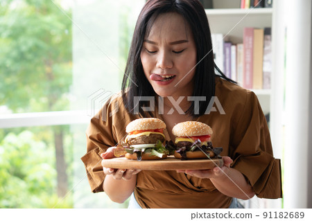 Hungry overweight woman holding hamburger on a wooden plate, During work from home, gain weight problem. Concept of binge eating disorder (BED). Hungry overweight woman holding hamburger on a wooden plate, During work from home, gain weight problem. Concept of binge eating disorder (BED). 91182689
