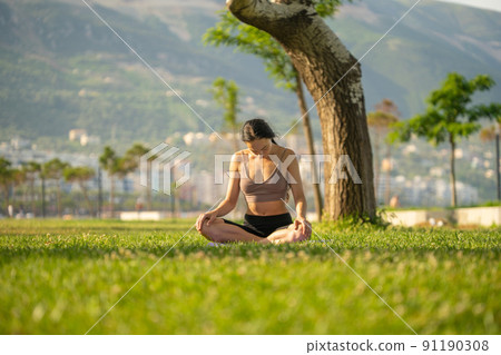 A woman in brown sport bra and black short sitting on yoga mat doing maditation with hand together at chest on grass floor and garden blurry background. A woman in brown sport bra and black short sitting on yoga mat doing maditation with hand together at chest on grass floor and garden blurry background. 91190308