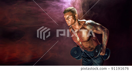 Brutal sweaty strong young man athlete with naked upper body standing doing workout with dumbbels and showing strong pumped up biceps over smoky background. Sport men body concept 91191676