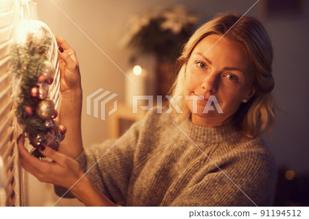 Portrait of content beautiful young woman in gray sweater standing in dark room illuminated with lamps and hanging Christmas wreath on door 91194512