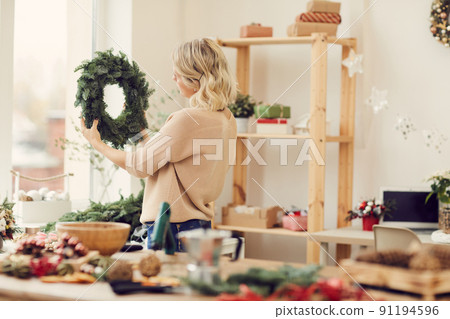 Blonde young woman in sweater standing in own workshop and looking at green Christmas wreath Blonde young woman in sweater standing in own workshop and looking at green Christmas wreath 91194596