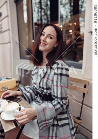 Pretty brunette woman sitting at table outdoors with cup of coffee and smiling. Cafe terrace. Pretty brunette woman sitting at table outdoors with cup of coffee and smiling. Cafe terrace. 91196098