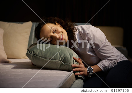 Beautiful relaxed Hispanic woman smiles toothy smile while putting her head on a green velour cushion sitting near a sofa bed with orthopedic mattress 91197196