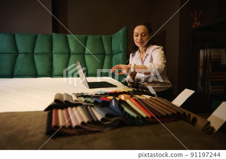 Creative interior designer checking time on her watch while working on laptop on orthopedic mattress next to laid out colorful fabric samples in home design studio Creative interior designer checking time on her watch while working on laptop on orthopedic mattress next to laid out colorful fabric samples in home design studio 91197244