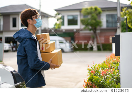 Asian postman with a hygiene protective face mask picking up a parcel package or parcel box from the truck for deliver to customer's door. New Normal concept during covid-19 or coronavirus pandemic Asian postman with a hygiene protective face mask picking up a parcel package or parcel box from the truck for deliver to customer's door. New Normal concept during covid-19 or coronavirus pandemic 91223675