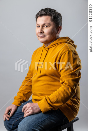 Portrait of attractive middle aged man looking at camera, sitting on chair and expressing positive emotions, wearing urban style hoodie. Indoor studio shot isolated on gray background. Portrait of attractive middle aged man looking at camera, sitting on chair and expressing positive emotions, wearing urban style hoodie. Indoor studio shot isolated on gray background. 91228550